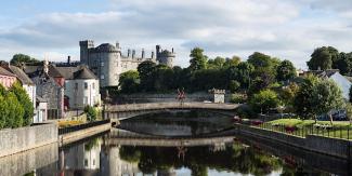 a photo of Kilkenny castle beside river, trees forming a backdrop to stone bridge