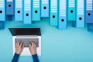 A series of blue ring binders arranged irregularly across the top; from below, a pair of hands typing on a silver laptop.