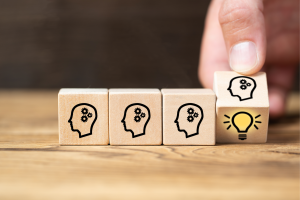 Lined up are three small wooden cubes with the engraved outline of a head with cog wheels; a hand is turning a fourth cube at the end of the sequence to reveal a yellow lightbulb, symbolising an idea.