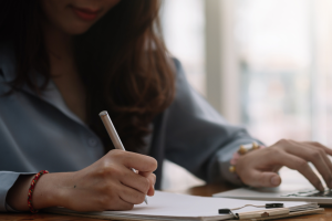 Young woman, partially out of frame, making notes and working on laptop, as if in preparation for a class or exam