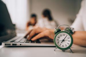 Hands typing on a laptop with small green alarm clock in foreground, indicating time pressure